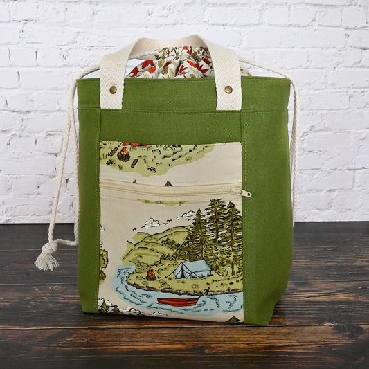 Fun green canvas project tote with lots of pockets.  Features a lovely picturesque cotton pocket on the front with a tenting scene by a lake and two exterior pockets.  The inside are made with green spotty and  leaf/acorn fabrics.  There are two interior pockets.  This bag features an expandable double drawstring is made in the leaf/acorn print.  This bag also has two convenient 18" handles in natural cotton webbing.  Made in Canada.